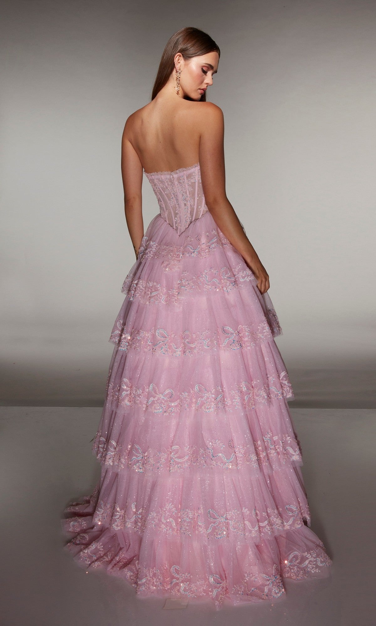 A woman faces away from the camera in the Alyce 62191 long prom dress, showcasing a strapless embellished corset bodice and tiered pink A-line skirt with lace and sequin details against a plain background.
